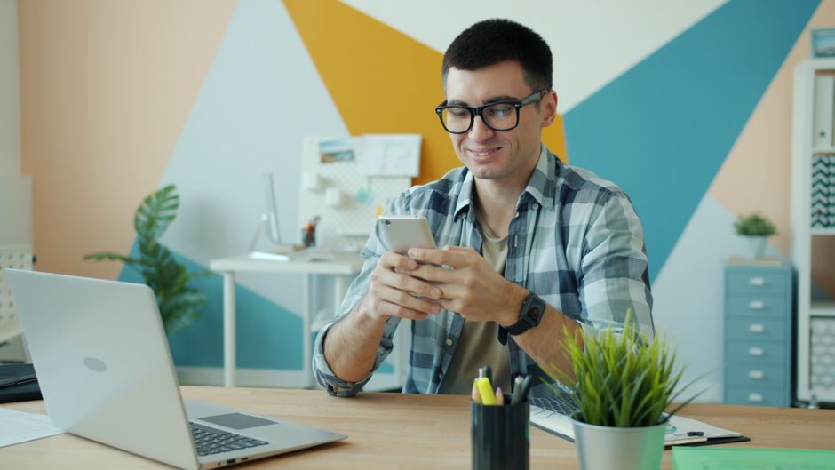 A young man in an office setting using a smartphone, with a laptop and stationery on the table.