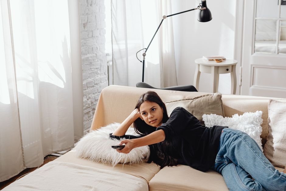 Young woman lounging on a cozy sofa, holding a remote control while relaxing at home.
